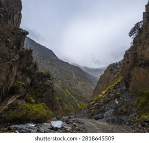 Wide-angle view of a rugged mountain gorge in Kyrgyzstan. Steep cliffs rise alongside a fast-flowing river and winding gravel road under an overcast sky.