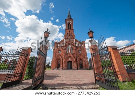 Wide-angle view through open wrought iron gates of the neogothic red brick church in Postavy, Belarus. Sunny sky with white clouds