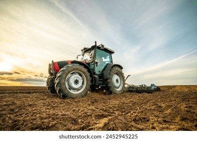 Wide-angle view of a tractor plowing farmland against a dramatic sunset sky - Powered by Shutterstock