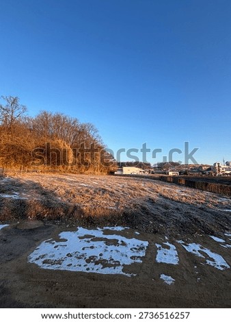 A wide-angle view of a vacant plot of land covered in withered grass and gravel, with a suburban residential neighborhood in the background.