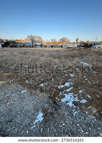 A wide-angle view of a vacant plot of land covered in withered grass and gravel, with a suburban residential neighborhood in the background.