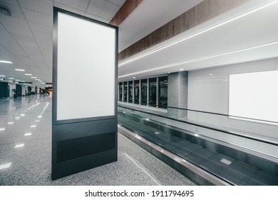 Wide-angle view of a vertical empty advertising billboard mockup next to the travelator indoors of a modern airport; a template of a blank information poster in the railroad terminal, moving walkway