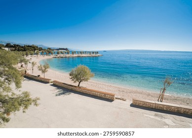 Wide-angle view of Znjan beach in Split, Croatia. Scenic coastline with stone benches, green trees, and crystal clear blue Adriatic sea under a bright sunny sky.