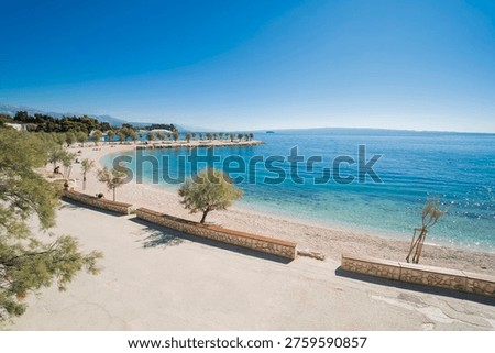 Wide-angle view of Znjan beach in Split, Croatia. Scenic coastline with stone benches, green trees, and crystal clear blue Adriatic sea under a bright sunny sky.