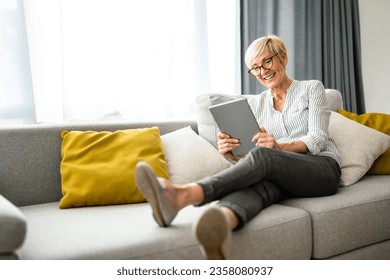 Widely smiling cheerful senior woman reading news on digital tablet while sitting on couch in living room. - Powered by Shutterstock