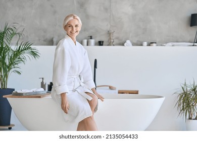 Wideshot Of Happy Smiling Beautiful Middle Aged Woman In Bathrobe At Luxury Spa Hotel Sitting On Bath Tube. Advertising Of Bodycare Spa Procedures Antiage Recreation Skin Care Salon Concept.