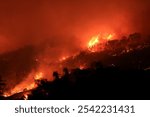  Wide-shot view of a ferocious wildfire spreading across a hillside, with intense flames engulfing the landscape.