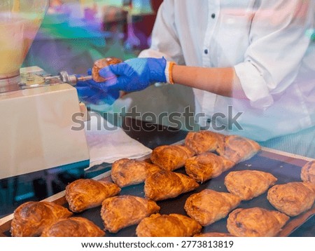 The widest angle pulls back to encompass the full bakery counter scene, with a staff member arranging golden turnovers against the backdrop of baking trays and display equipment. This establishing shot places the apple turnovers in their complete commercial context, from production tray to retail display, capturing the full lifecycle of a bakery product in one frame.