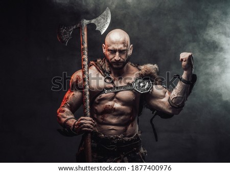 Wielding an axe and dressed in light armour with fur bald and furious viking warrior poses in dark and smokey background showing his huge biceps.
