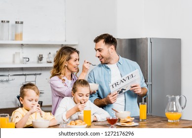 Wife Feeding Husband Porridge With Spoon While Children Eating Oatmeal At Table