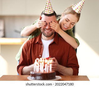 Wife Making Surprise For Husband,  Covering His Eyes With Hands Excited Man Holding Gift And Being Happy While Sitting In Kitchen At Table With Cake. Family Couple In Party Hats Celebrating Birthday