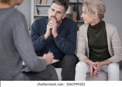Wife supporting her husband in therapy with the man listening curiously to the counselor in a gray office