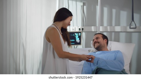 Wife Visiting And Talking With Patient Husband In Hospital Bed. Portrait Of Young Woman Visitor Smiling And Holding Hand Of Sick Adult Brother Resting In Bed At Hospital Ward