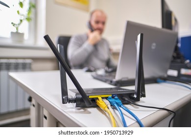 Wi-Fi router mounted on the edge of the table with a lot of antennas and cables. In the background, a man is talking on the phone. The router is installed in the office.