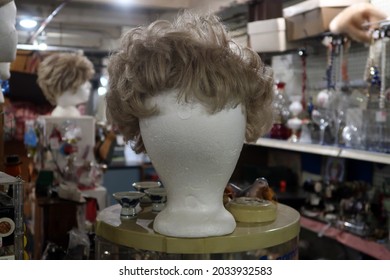 Wig Stand With A Used Wig On In In The Foreground. Miscellaneous Merchandise In The Background. 