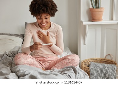 It Wil Be Start Of New Life! Cheerful African American Future Mother Cant Believe In Positive Result On Pregnancy Test, Awakes Early In Morning, Wears Casual Pyjamas, Poses Against Bedroom Interior