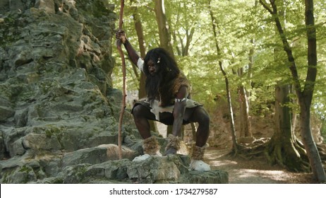 Wild African Savage In Animal Fur Costume Sitting On Rocks Watching Animals Food Hunting In The Forest Near Cave. Tribe Leader. Homo Sapiens.