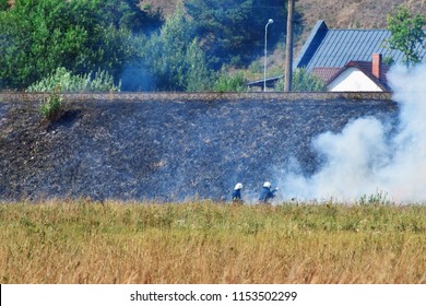 Wild Fire And Smoke In Dry Meadow Grass Due To Hot Windy Weather In Summer Started To Burn From Railway Behind And Two Firefighters Wearing  Helmets Working. Firefighting Rescue Operation Concept.
