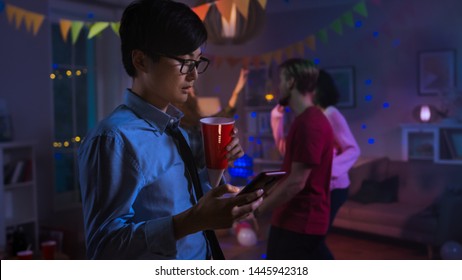 At The Wild House Party: Confident Asian Man Uses Smartphone Instead Of Dancing With Other People.