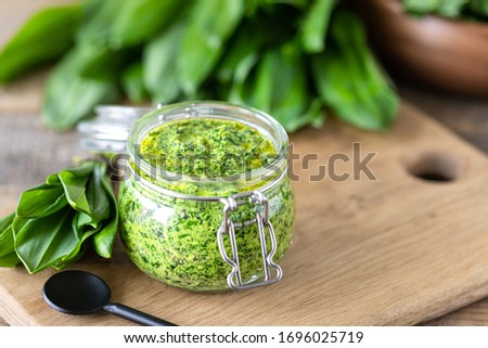 Wild leek pesto with olive oil and parmesan cheese in a glass jar on a wooden table. Useful properties of ramson. Leaves of fresh ramson.
