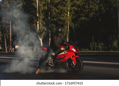 Wild motorcyclist in black helmet and leather jacket lets tires spin during burnout and making smoke. Young brave stuntman sitting on red sports motorcycle doing the trick while looking at camera