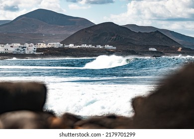 Wild rocky coastline of surf spot La Santa Lanzarote, Canary Islands, Spain. Surfer riding a big wave in rocky bay, volcano mountain in background.