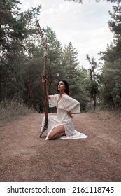 wild shaman party. young brunette girl in white boho style dress is sitting with long high wooden witch staff in hand and looking apart on a pine forest background. lifestyle concept, free space