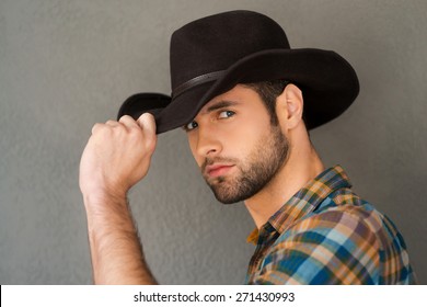 Wild West is his heart. Handsome young man adjusting his cowboy hat and looking at camera while standing against grey background 