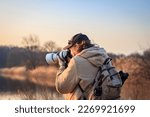 Wildlife photographer with camera photographing bird on lake at sunset. Man with backpack hiking in nature