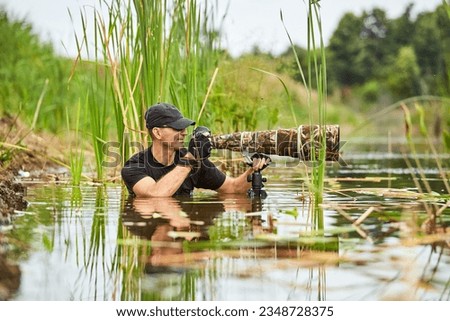Wildlife photographer outdoor, standing in the water. Wildlife photographer in summer time working in the wild. Wildlife photographer using telephoto lens with camouflage coating