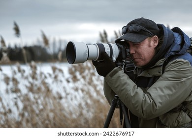 Wildlife photographer is setting up camera on tripod outdoors. Man photographing landscape or animals at lake in winter. Bird watching - Powered by Shutterstock