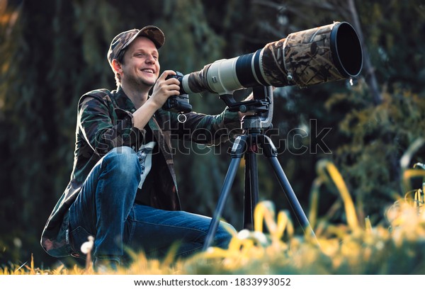 wildlife photographer using telephoto lens with\
camouflage coating photographing wild life using gimbal head on\
tripod. professional photography equipment for cinematic shooting\
in the nature outdoor