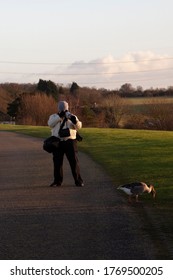 A Wildlife Photographer Wearing Winter Jacket, Cap And Fingerless Gloves Is Focusing On A Goose Using A Telephoto Lens. He Is Well Equiped With Camera Bags And Binoculars