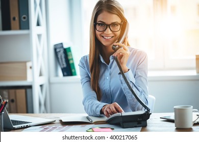 I will connect you in one second! Cheerful young beautiful woman in glasses talking on the phone and looking at camera with smile while sitting at her working place