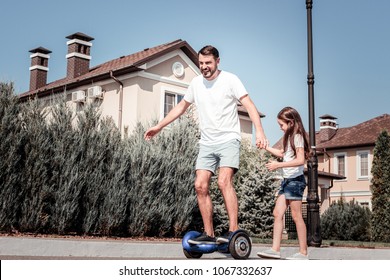 I Will Help You. Amazed Pleasant Unconfident Man Standing On The Self-balancing Scooter Holding Daughters Hand And Smiling.