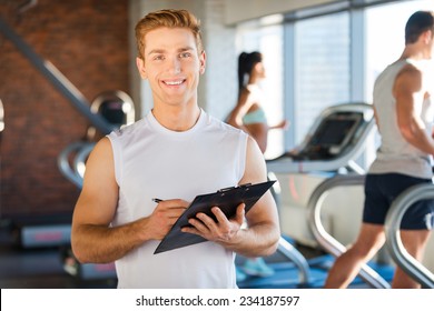 I will make you healthy! Handsome young man holding clipboard and smiling while people running on treadmill in the background   - Powered by Shutterstock