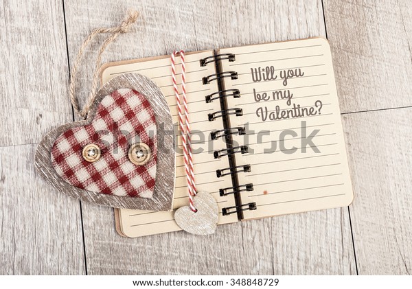 Will you be my\
Valentine Text in open lined notebook next to wooden-textile heart\
and book divider. Valentine day romantic greeting card design. This\
image is toned