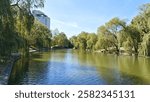 Willows and other deciduous trees grow on the grassy concreted banks of the canal, and buildings stand. A pedestrian walkway with a stone parapet runs nearby. Sunny autumn weather and blue sky