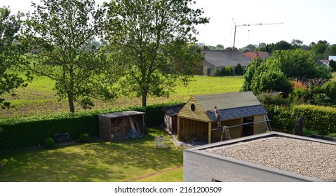 Wilsele, Vlaams-Brabant, Belgium - May 27, 2022:neighbor Stands Dangerously Unstable On Wooden Planks Supported By A Ladder And Scaffolding Beam, Completes His Work On The Roof  Nailing Roof Shingles