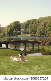 Wiltshire, UK. May 7 2018. A Couple Enjoy The Early May Bank Holiday Record Breaking High Temperatures By Relaxing In The Grass Next To A Lake.            