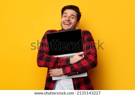 Win Concept. Portrait of overjoyed excited young man hugging notebook with black empty screen, holding it tight near chest. Positive male nerd posing isolated over yellow studio background wall