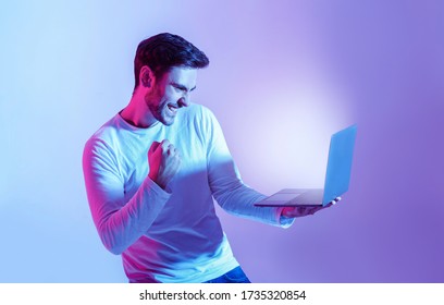 Win online. Man expresses emotions of happiness, looking in laptop with luminous screen, isolated in neon, studio shot