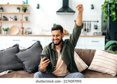 Win, Success. Indian Cheerful Happy Young Indian Or Arabian Man In Casual Wear, Sit At Home In Living Room On Comfortable Sofa, Using Smart Phone, Read Good News, Gesturing With Hand, Smiling Joyfully