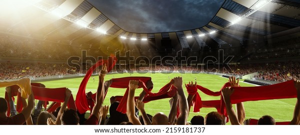 Win, victory of favourite team. Back view of\
football, soccer fans cheering their team with red scarfs at\
crowded stadium at evening time. Concept of sport, cup, world,\
event, competition
