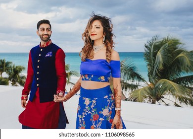 Wind blows around Hindu groom in blue sherwani and bride in lehenga posing in white house with gorgeous seaside view behind them