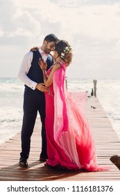 Wind blows bride's pink sari while she stands with Hindu groom on a wooden quay among foaming waves