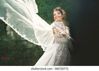 Wind blows bride's veil while she poses before the wall covered with green ivy and red flowers