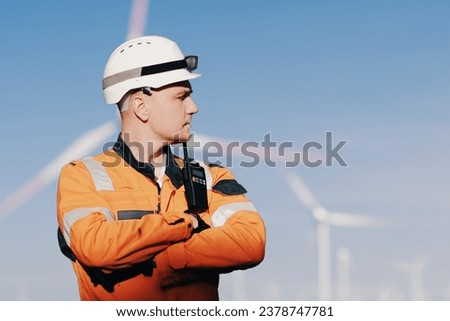 Wind Farm Offshore Maintenance Technician. Seafarer. Seaman. Navigator. A Man In A Working Overall Boiler Suit With A Radio And Safety Helmet With A Blurred Wing Generators In The Background