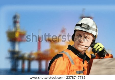 Wind Farm Offshore Maintenance Technician. Seafarer. Seaman. Navigator. A Man In A Working Overall Boiler Suit With A Radio And Safety Helmet With A Blurred Oil Rig Offshore Platform In The Background