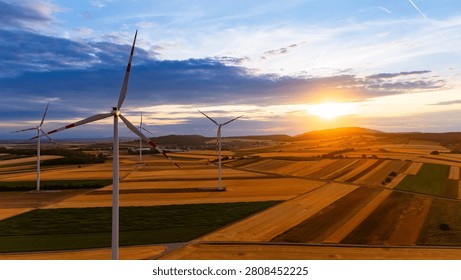 Wind farm over fields with setting sun. Oblique drone view, foreground turbine, roads, hills.